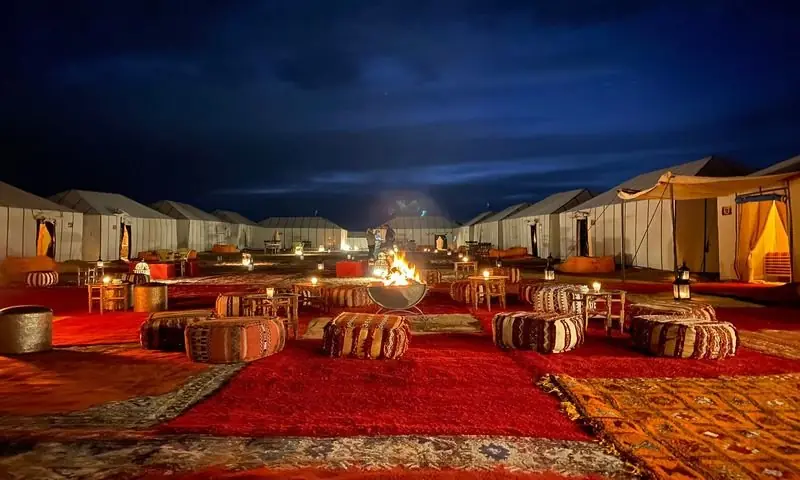 Acampamento no deserto à noite, com tapetes, pufes e fogueira entre as tendas